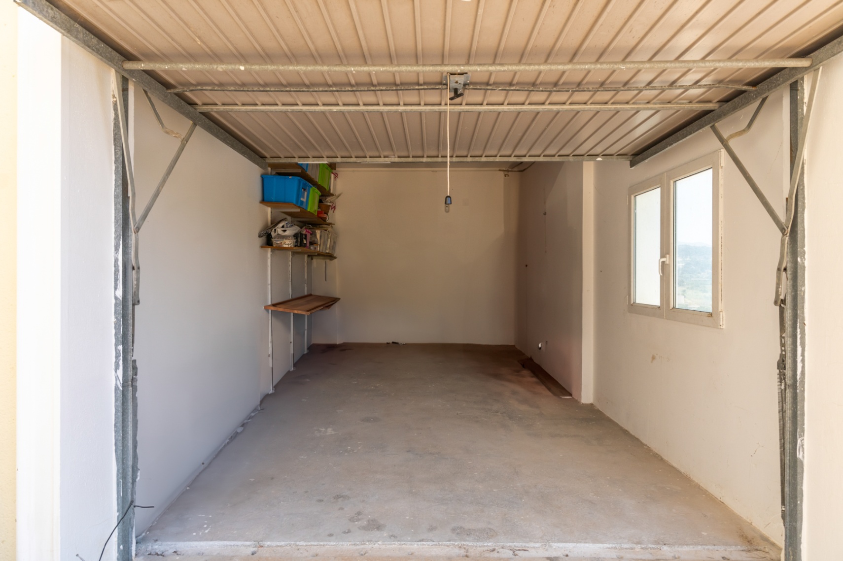 Garaje privado con suelo de hormigón, paredes blancas y ventana lateral. Incluye estanterías de almacenamiento y puerta metálica basculante.