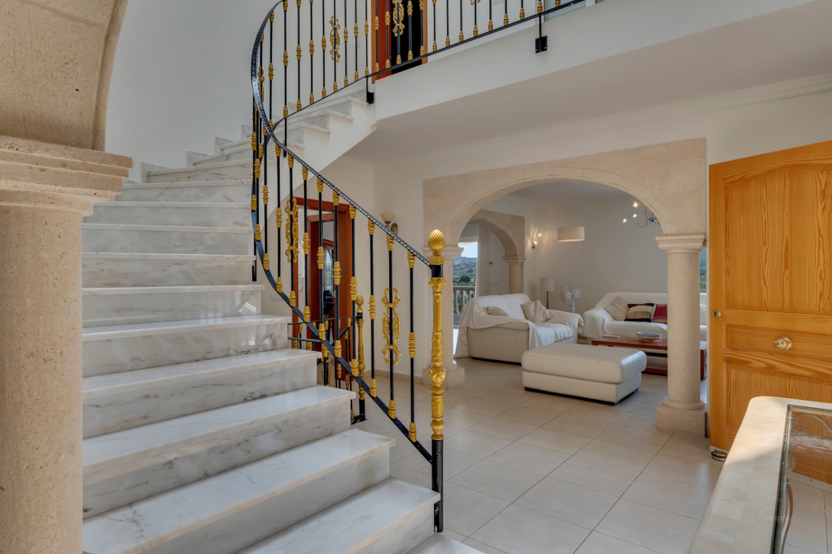 Hall de entrada con escalera de mármol y barandilla de forja ornamental. Arcos de piedra natural conectan con el salón, destacando los suelos cerámicos y la carpintería de madera.