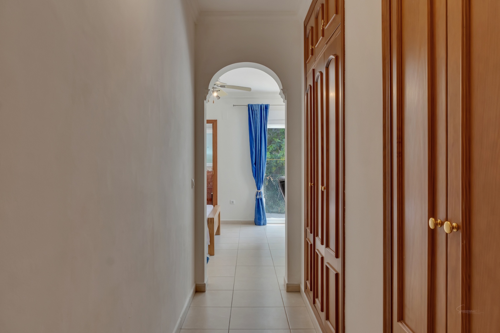 Pasillo con armarios empotrados de madera y puertas con molduras que conduce a un dormitorio a través de un arco decorativo, con suelos cerámicos claros.