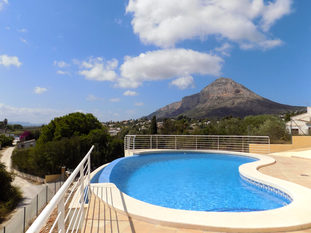 Piscina de forma curva con borde infinito y vistas panorámicas directas al macizo del Montgó, rodeada de una terraza pavimentada y barandillas de seguridad blancas.