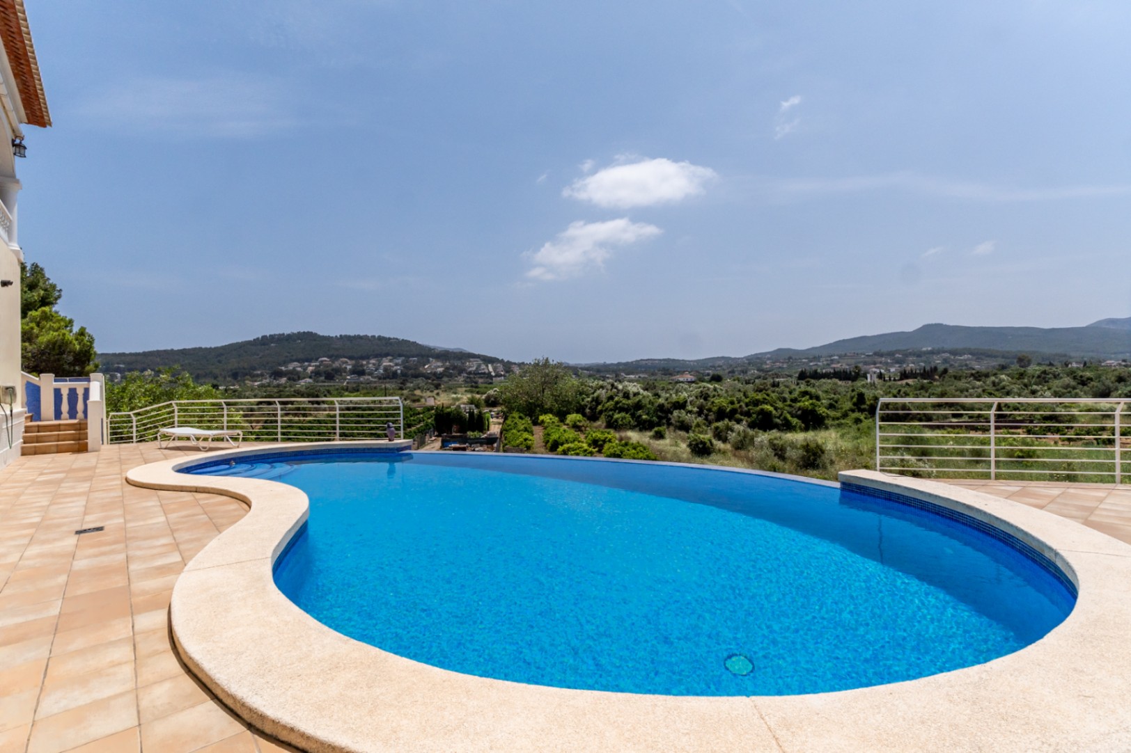Piscina infinity de diseño curvo rodeada por una terraza de baldosas cerámicas, con vistas panorámicas despejadas hacia el valle y las montañas de Jávea.