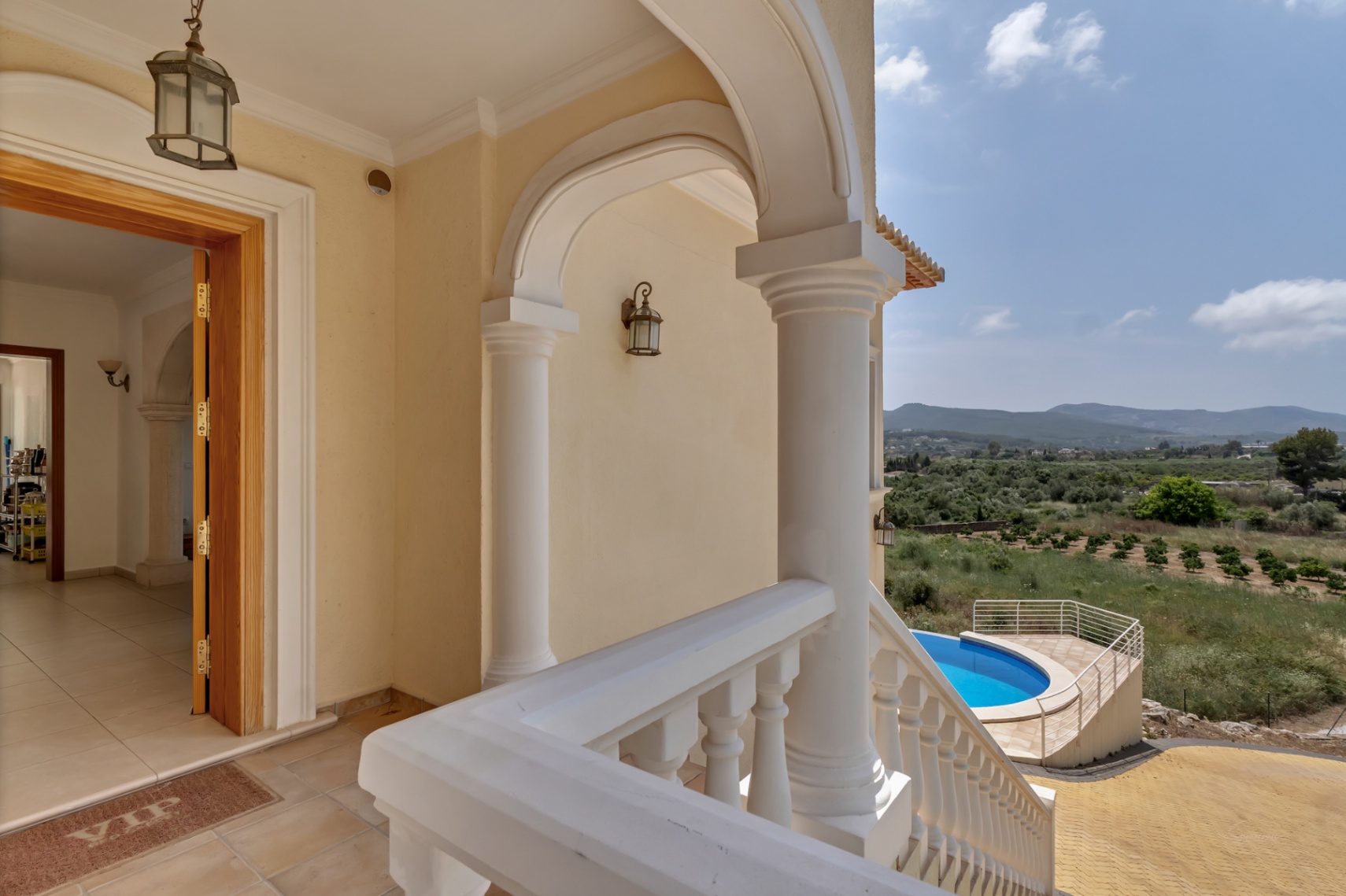 Porche de entrada con columnas blancas y balaustrada, ofreciendo vistas elevadas a la piscina circular y al valle circundante bajo un cielo despejado.