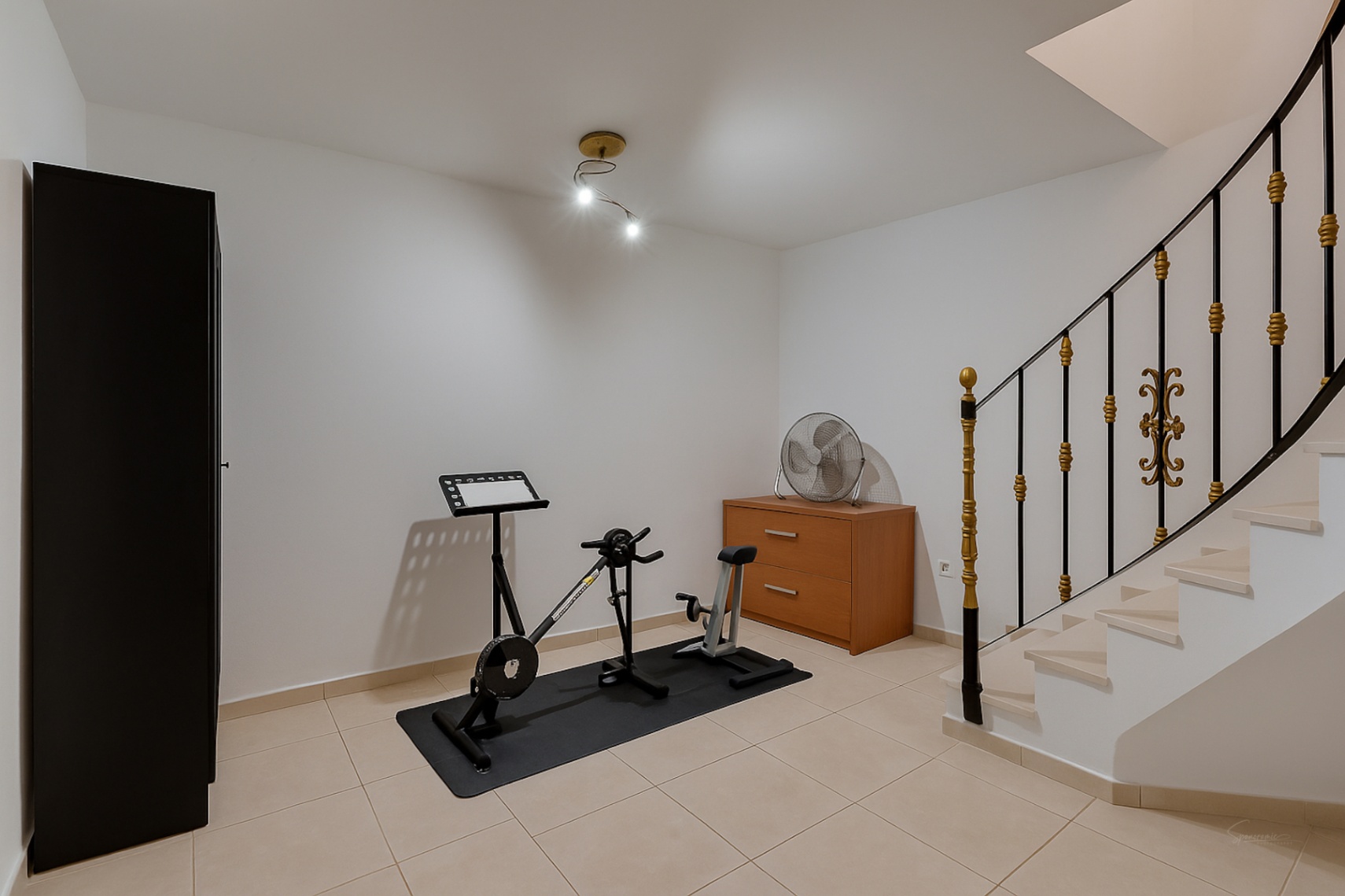 Sala polivalente en la planta inferior equipada con bicicleta estática, armario de almacenamiento y escalera con barandilla de hierro forjado y detalles dorados.