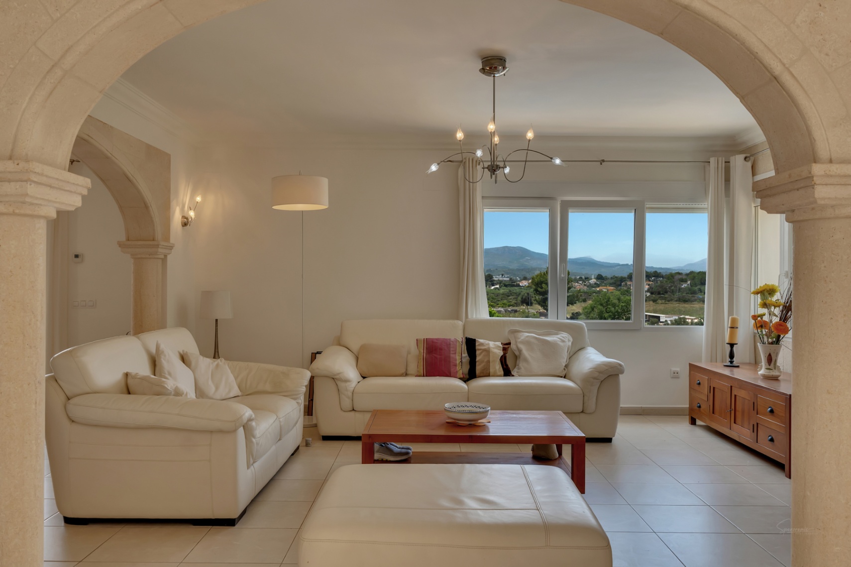 Salón enmarcado por un arco de piedra tosca, con sofás de cuero blanco y vistas panorámicas al valle y las montañas a través del ventanal principal.