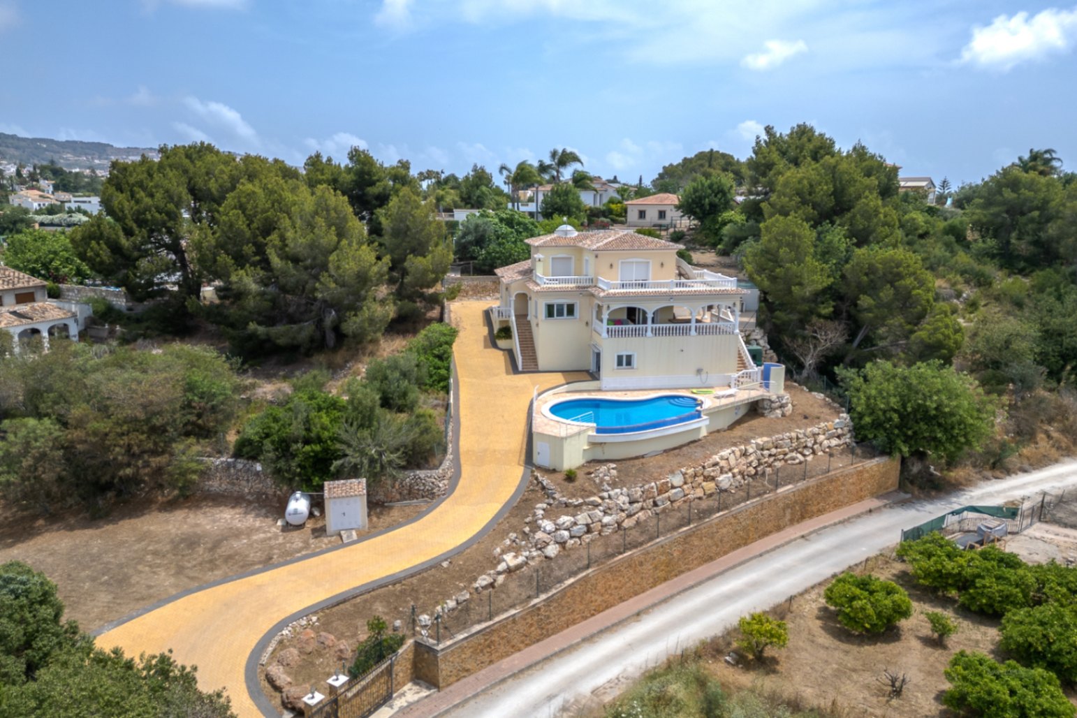 Vista aérea de la villa con piscina infinity, amplias terrazas y un camino de acceso privado rodeado de vegetación mediterránea y muros de piedra natural.