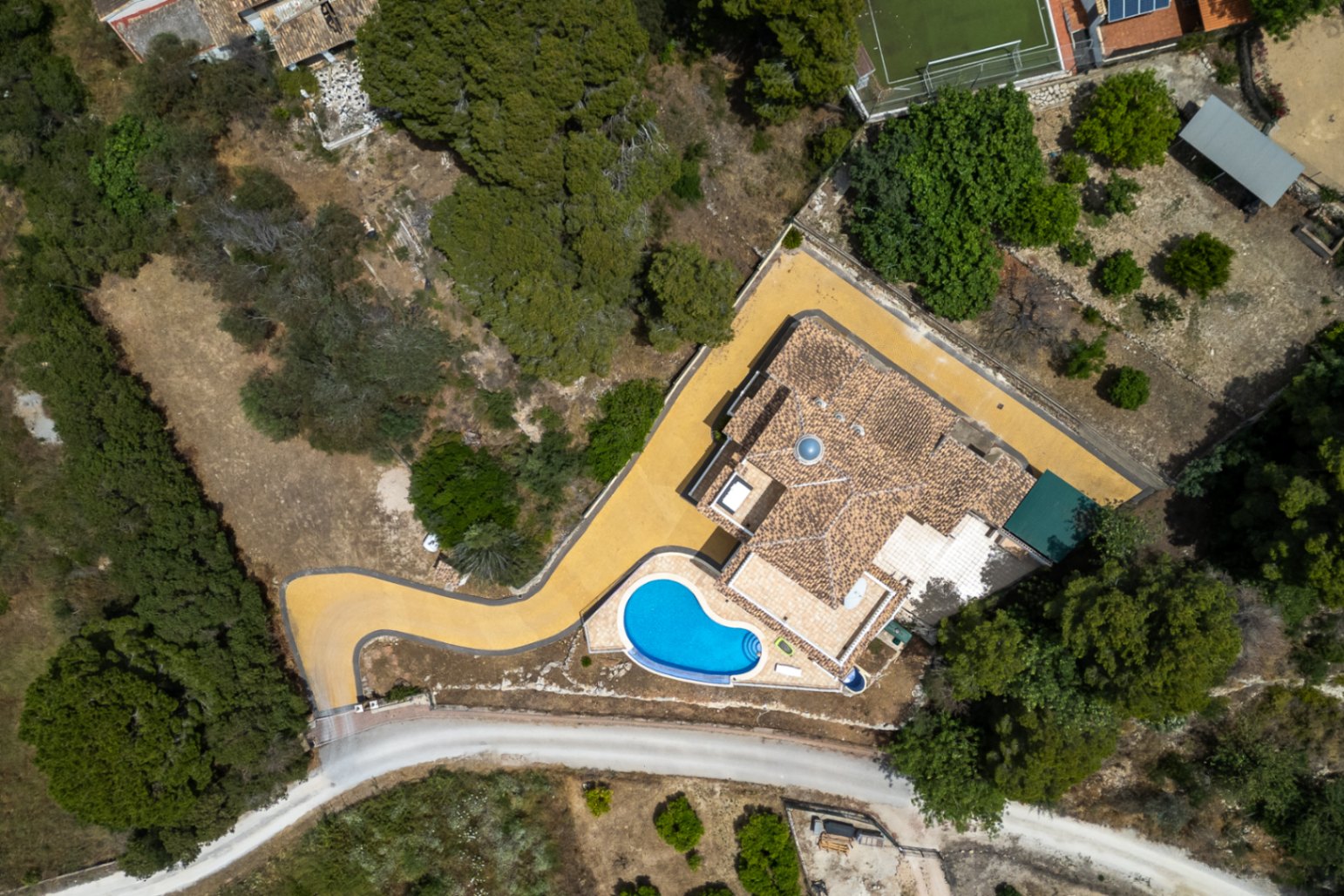 Vista aérea de la villa que muestra el tejado de teja con cúpula central, la piscina de forma orgánica y el acceso pavimentado rodeado de vegetación mediterránea.