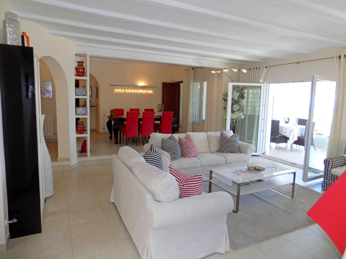 Amplio salón comedor con techos de vigas blancas, mobiliario en tonos neutros y acceso directo a una luminosa terraza acristalada. Al fondo destaca el comedor con sillas rojas.