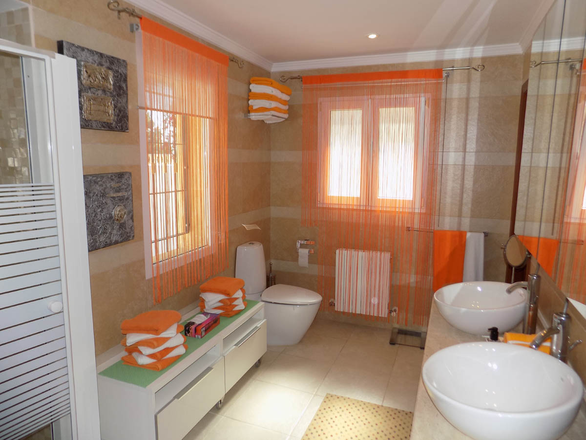 Baño completo con lavabo de doble seno sobre encimera, paredes alicatadas en tonos crema y ventanas con luz natural. Incluye radiador y sanitarios de diseño moderno.