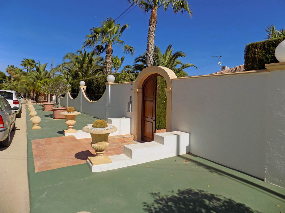 Entrada peatonal con puerta de madera en arco, muro perimetral blanco decorado con maceteros de piedra y palmeras mediterráneas.