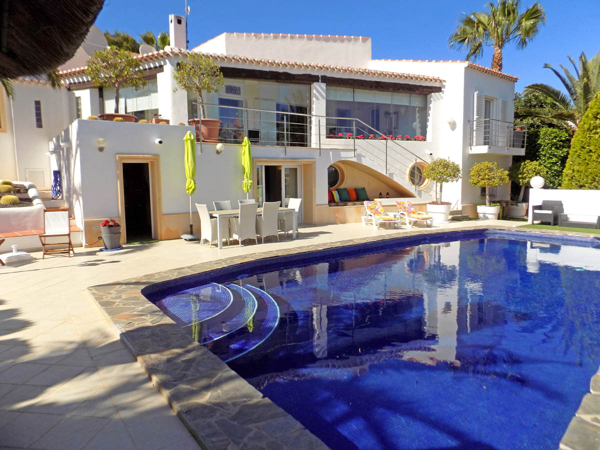Exterior de la villa con amplia piscina de mosaico azul, zona de comedor al aire libre y terraza acristalada en la planta superior rodeada de vegetación mediterránea.
