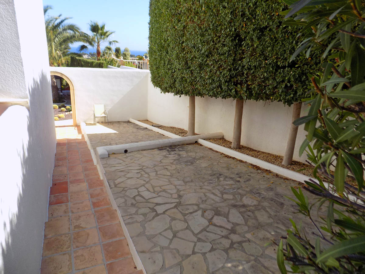 Patio exterior con pavimento de piedra natural y muros blancos, delimitado por setos altos. Un camino de baldosas de barro conduce a un arco con vistas a las palmeras.