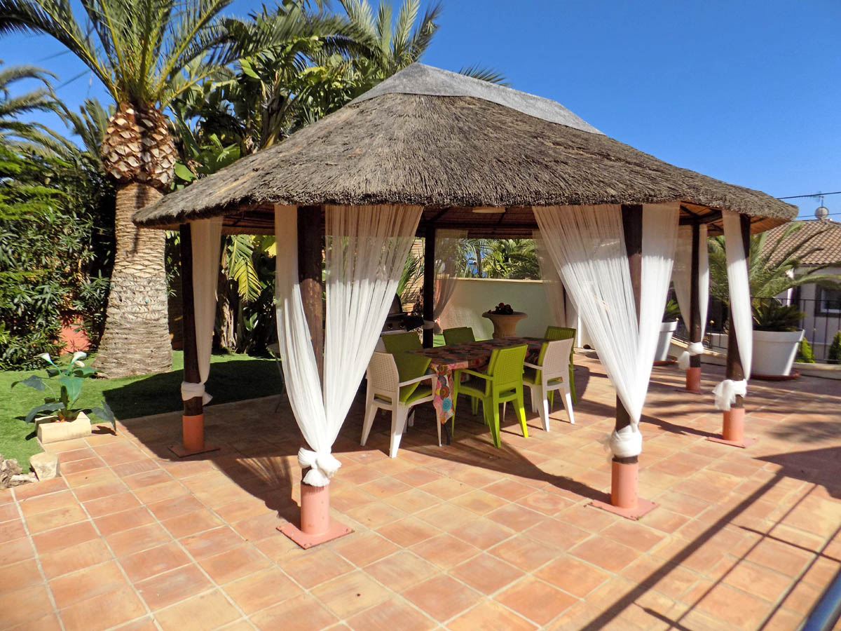 Pérgola exterior con techo de brezo y cortinas blancas sobre suelo de terracota, integrada en un jardín con palmeras y zona de comedor.