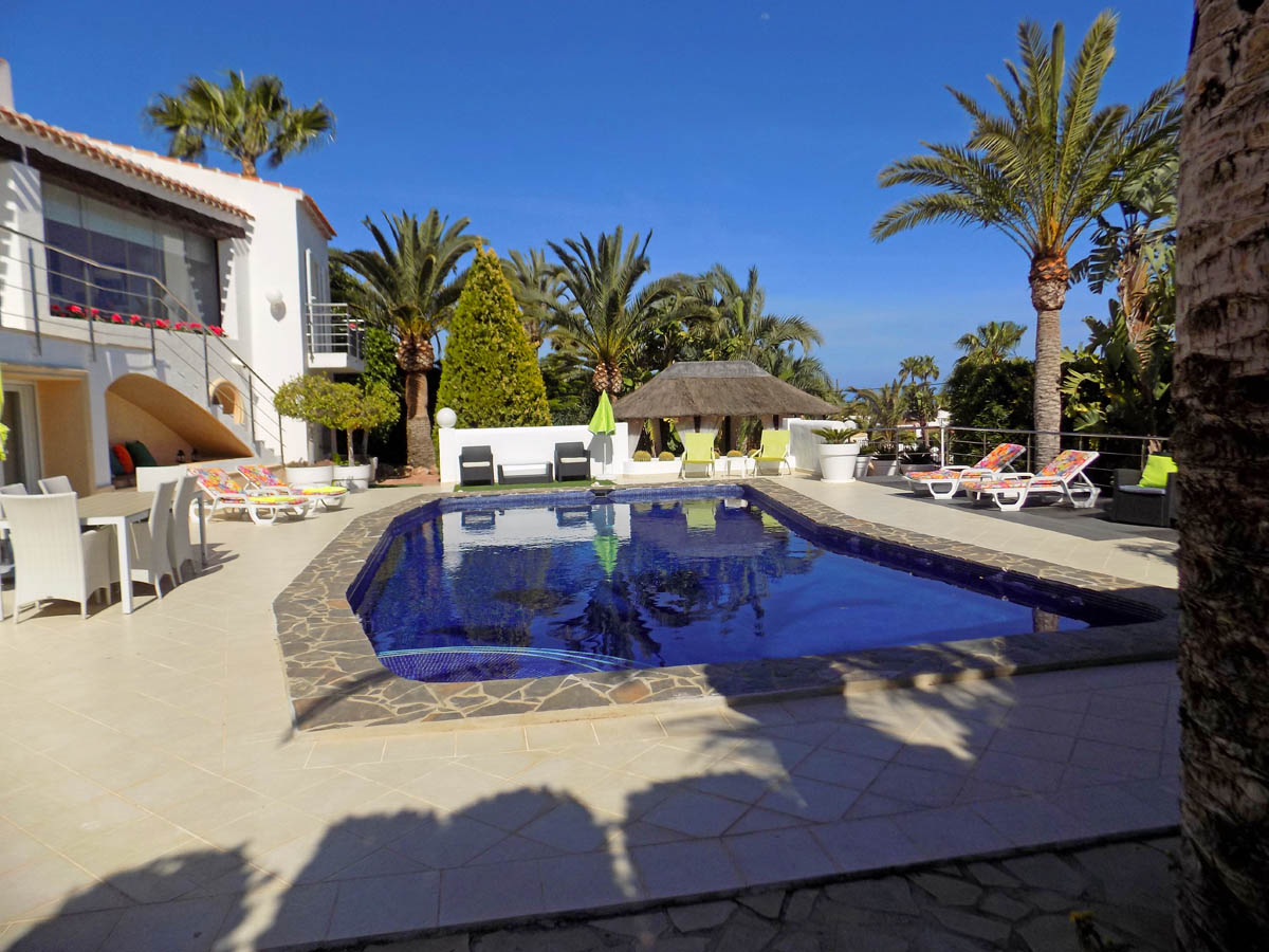 Piscina exterior con borde de piedra, amplia terraza con zona de comedor, tumbonas y una cabaña chill-out rodeada de palmeras y vegetación.