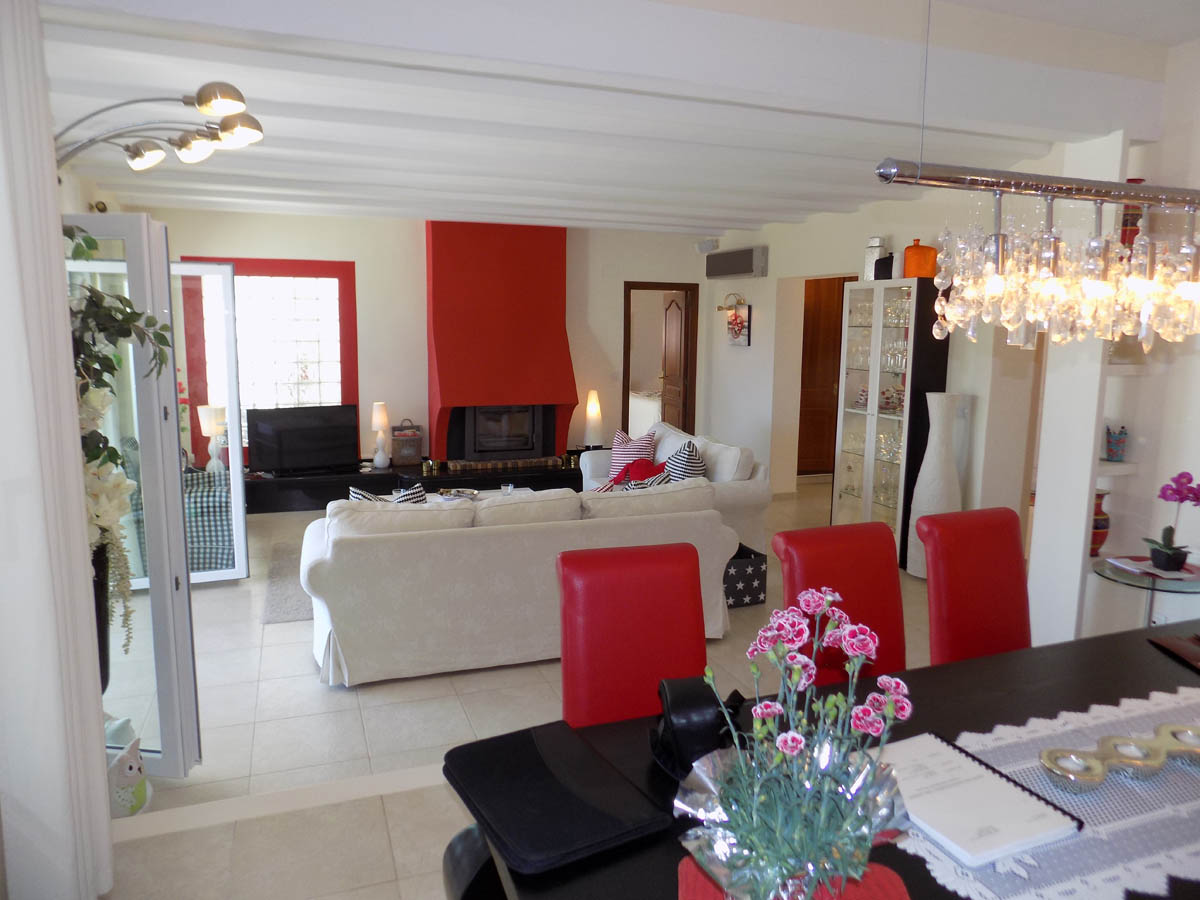 Salón comedor de planta abierta con una chimenea roja distintiva, techos con vigas blancas y mobiliario en tonos neutros con acentos rojos vibrantes.