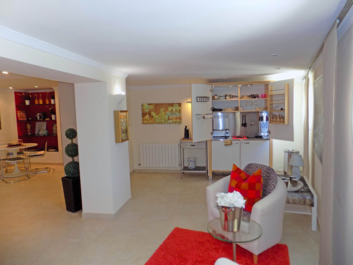 Salón en planta baja con cocina compacta integrada, zona de estar con sillón blanco y alfombra roja, y comedor al fondo con estanterías decorativas sobre pared roja.
