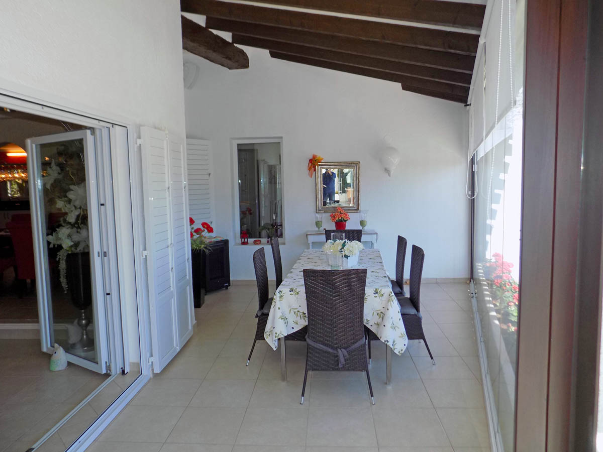 Terraza acristalada con vigas de madera a la vista, comedor para seis comensales y acceso directo al interior mediante puertas correderas blancas.