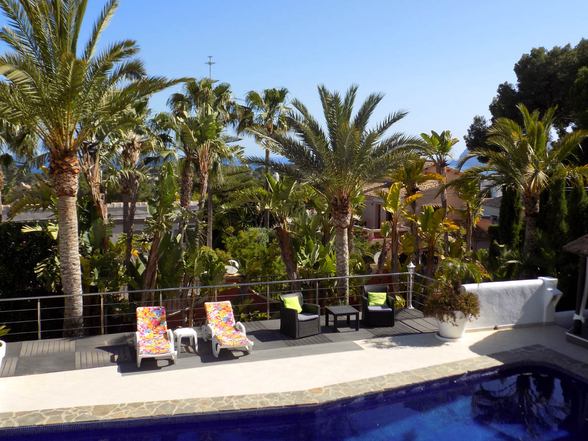 Terraza elevada con piscina y zona de solárium con tarima de madera, rodeada de palmeras maduras y vistas parciales al mar Mediterráneo.