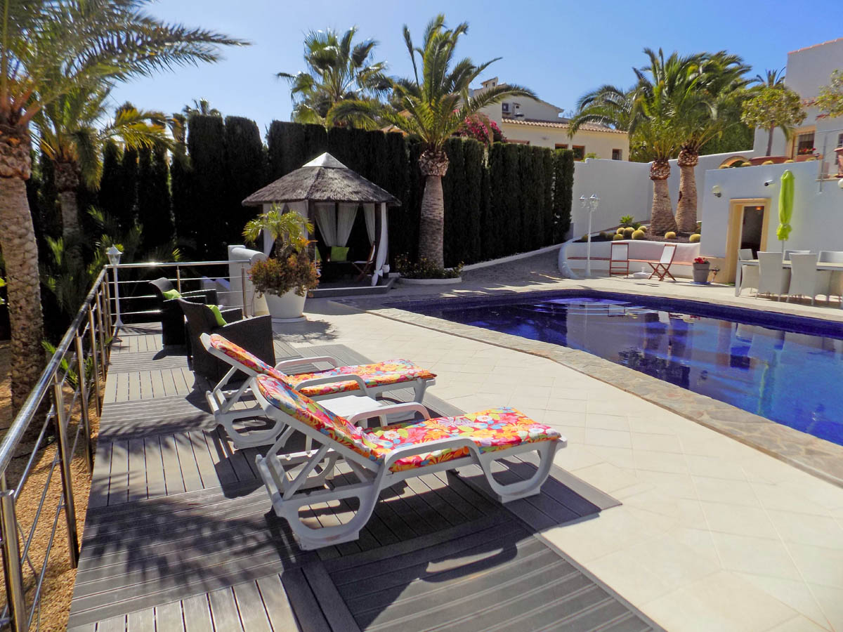 Zona de piscina con solárium de gres y madera tecnológica, tumbonas, cenador balinés con techo de brezo y vegetación mediterránea perimetral.