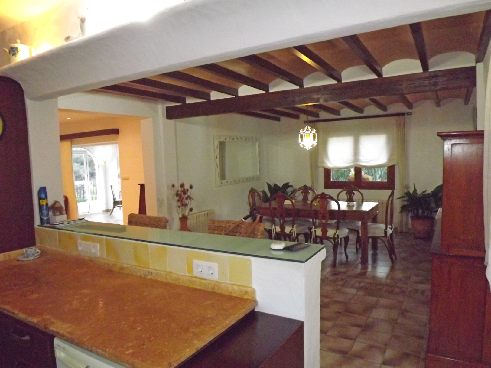 Comedor de estilo rústico con vigas de madera vistas y bovedillas, conectado a la cocina mediante una barra con encimera de piedra y estante de cristal.