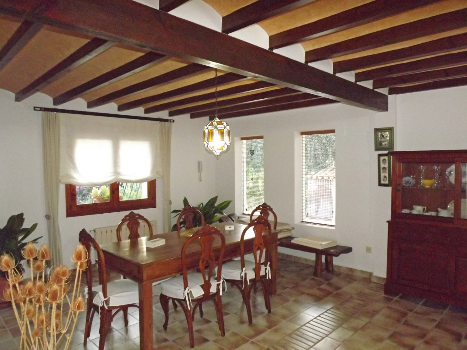 Comedor rústico con vigas de madera a la vista, mesa de madera maciza con sillas talladas y suelos de gres. Grandes ventanales aportan luz natural al espacio.