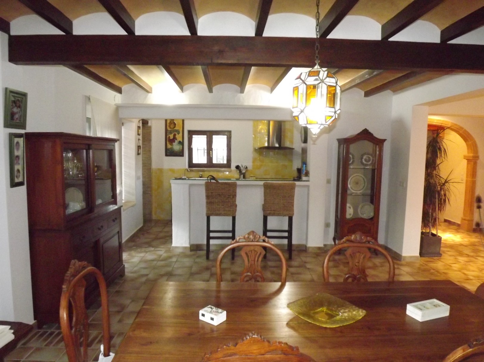 Comedor rústico con vigas de madera vistas, mesa de madera maciza y cocina abierta con barra americana y taburetes de mimbre bajo un techo de bovedillas.