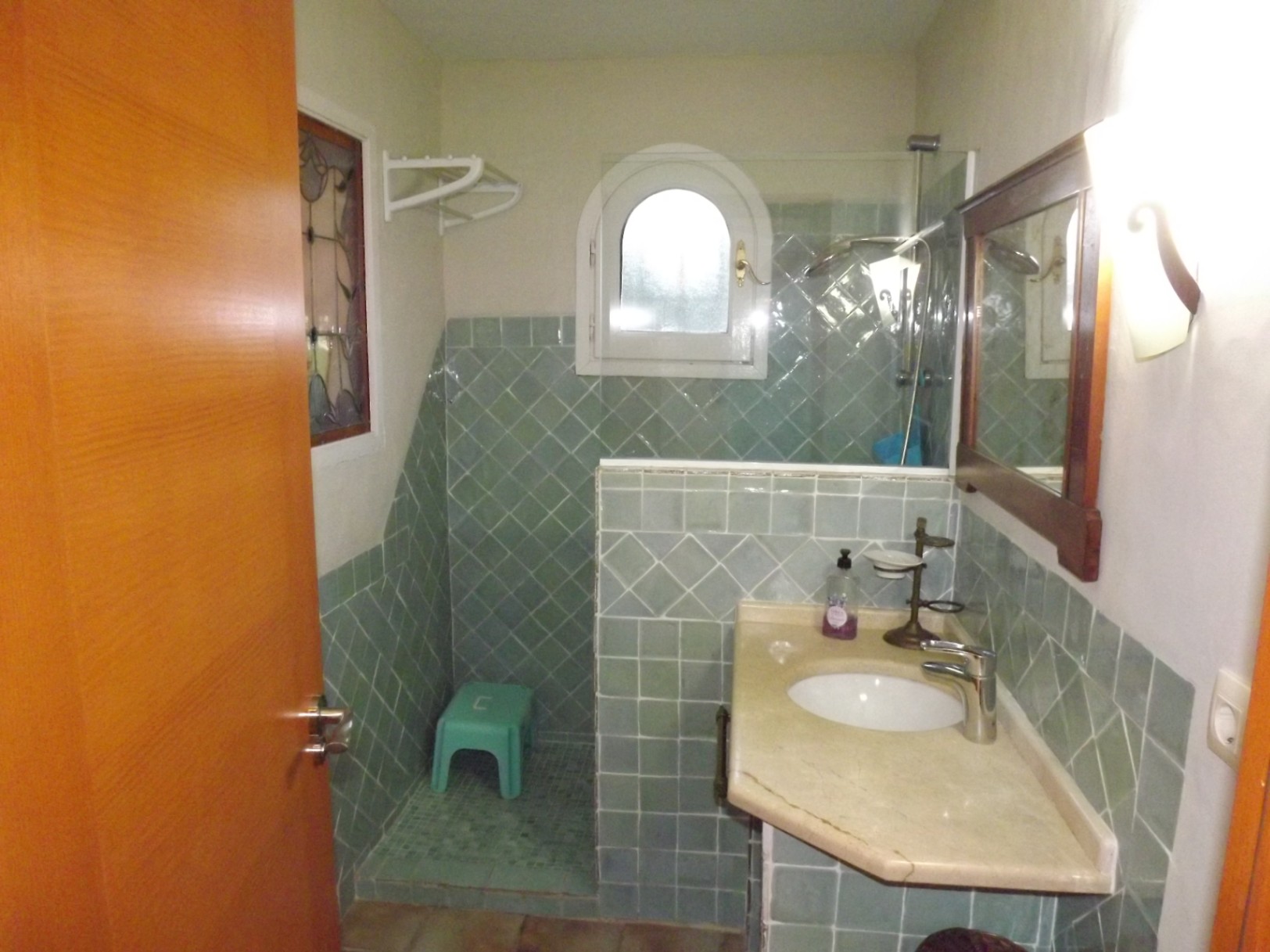 Cuarto de baño con azulejos verdes, encimera de piedra natural y ducha de obra con ventana de arco y mampara de cristal. Incluye un detalle de vidriera decorativa.