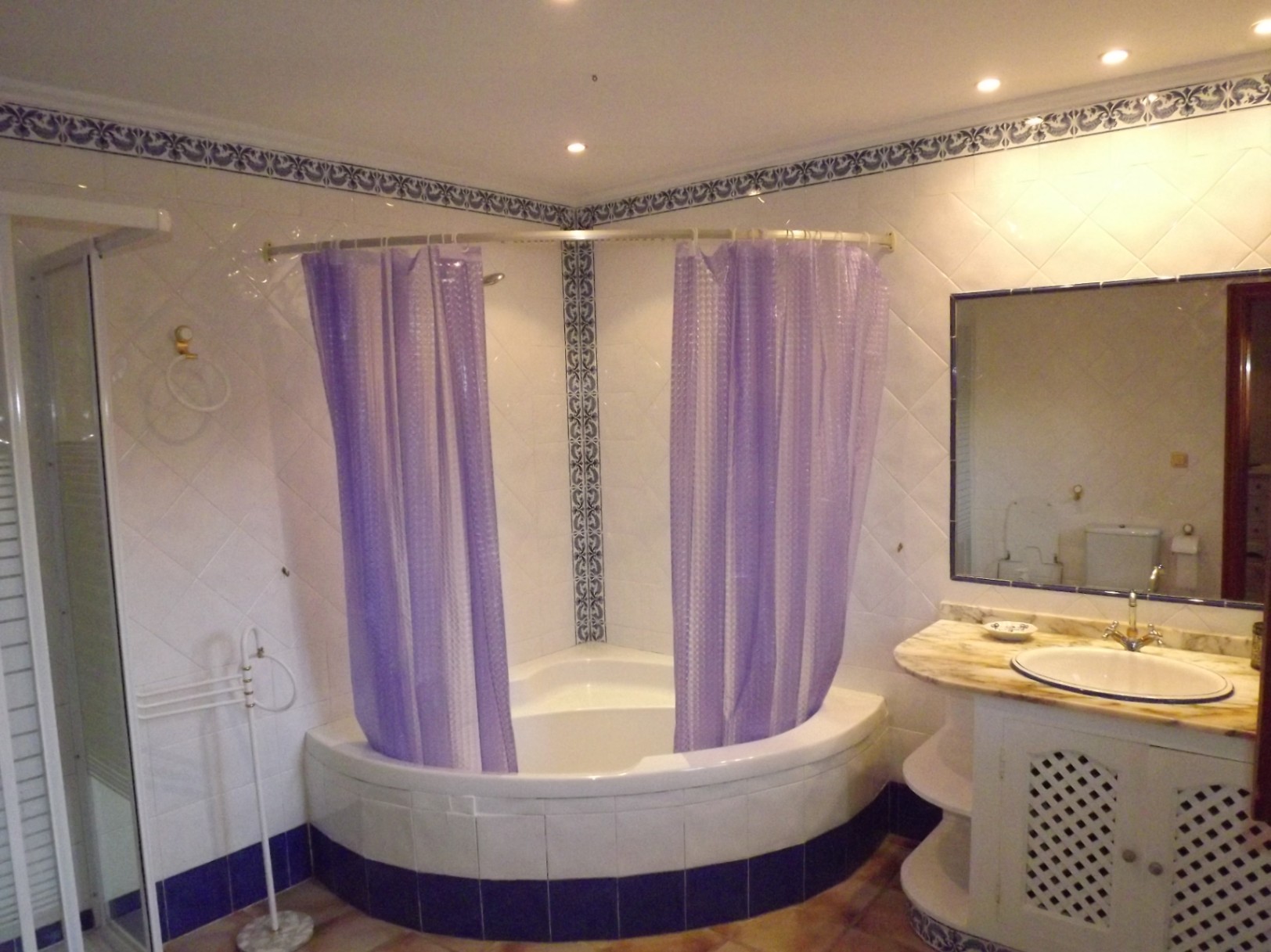 Cuarto de baño con bañera de esquina, cortinas púrpuras y azulejos blancos con cenefas azules. Incluye mueble de lavabo con encimera de mármol y espejo de gran formato.