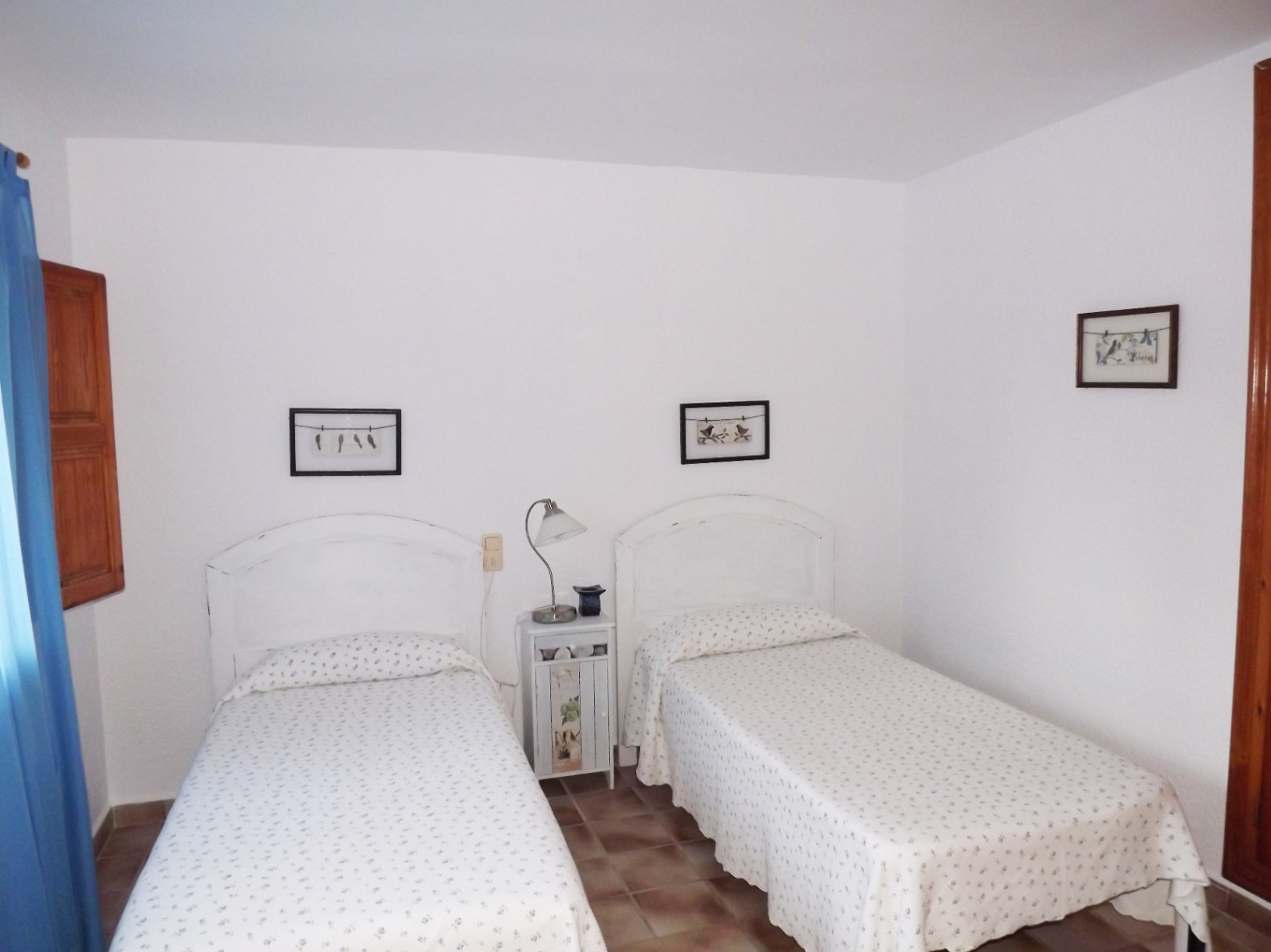 Dormitorio con dos camas individuales, cabeceros de madera blanca y suelos de baldosa cerámica. Incluye mesita de noche central y ventana con contraventana de madera.
