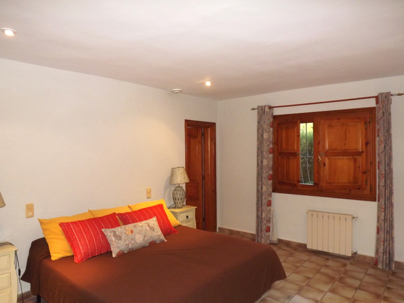 Dormitorio doble con suelos de gres, ventana de madera con contraventanas y calefacción central. Incluye iluminación empotrada y mobiliario rústico.