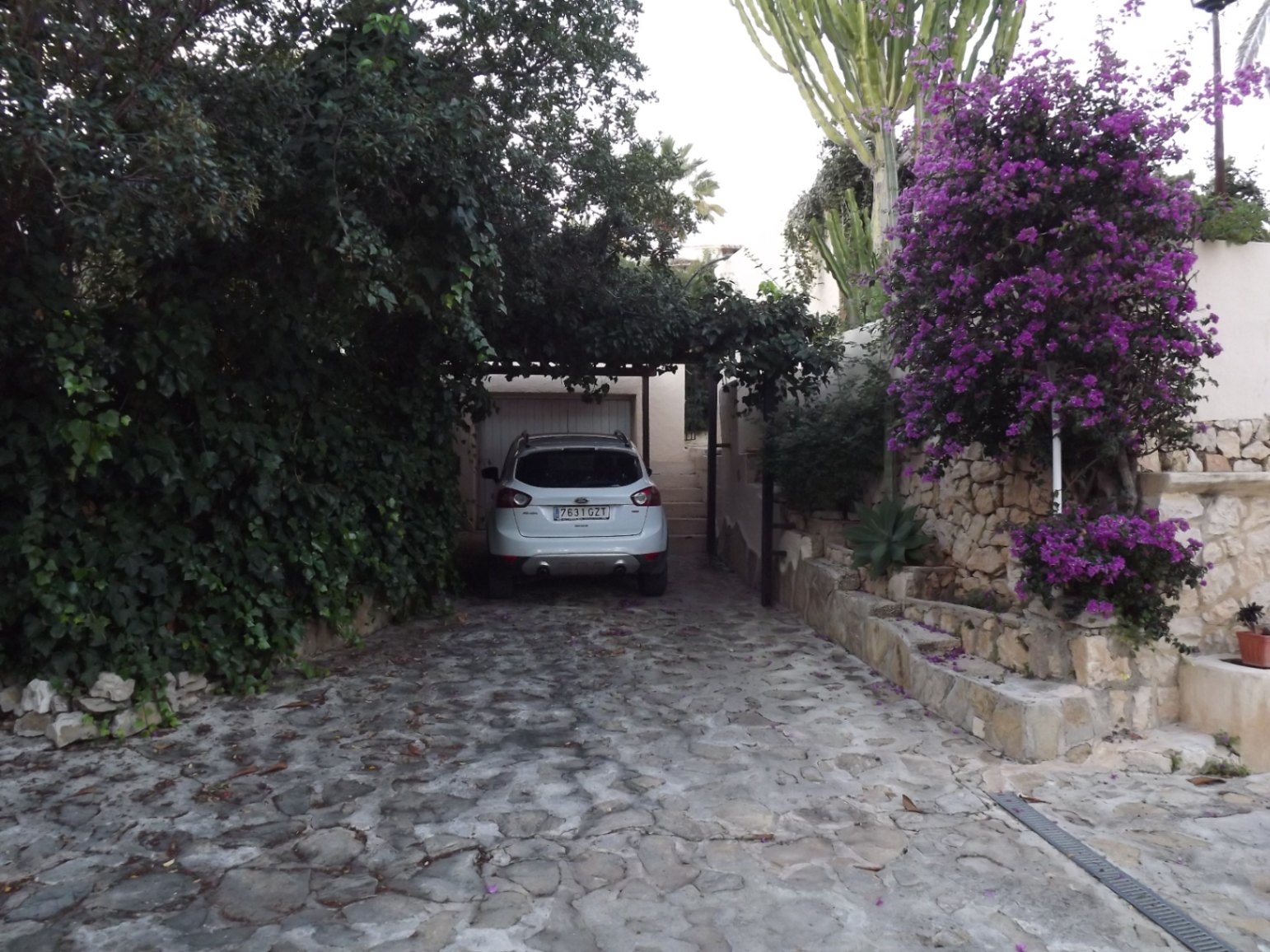 Entrada de piedra natural con acceso al garaje, rodeada de vegetación mediterránea, buganvillas y muros de piedra seca tradicionales.