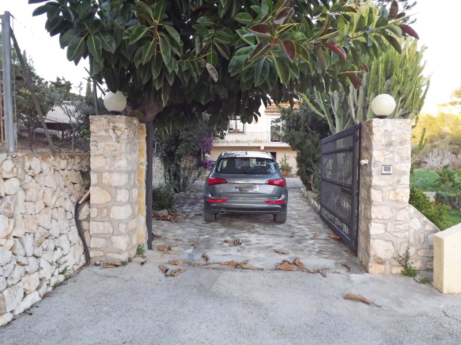 Entrada principal con pilares de piedra natural, iluminación esférica y portón metálico que da paso a un camino pavimentado rodeado de vegetación mediterránea.
