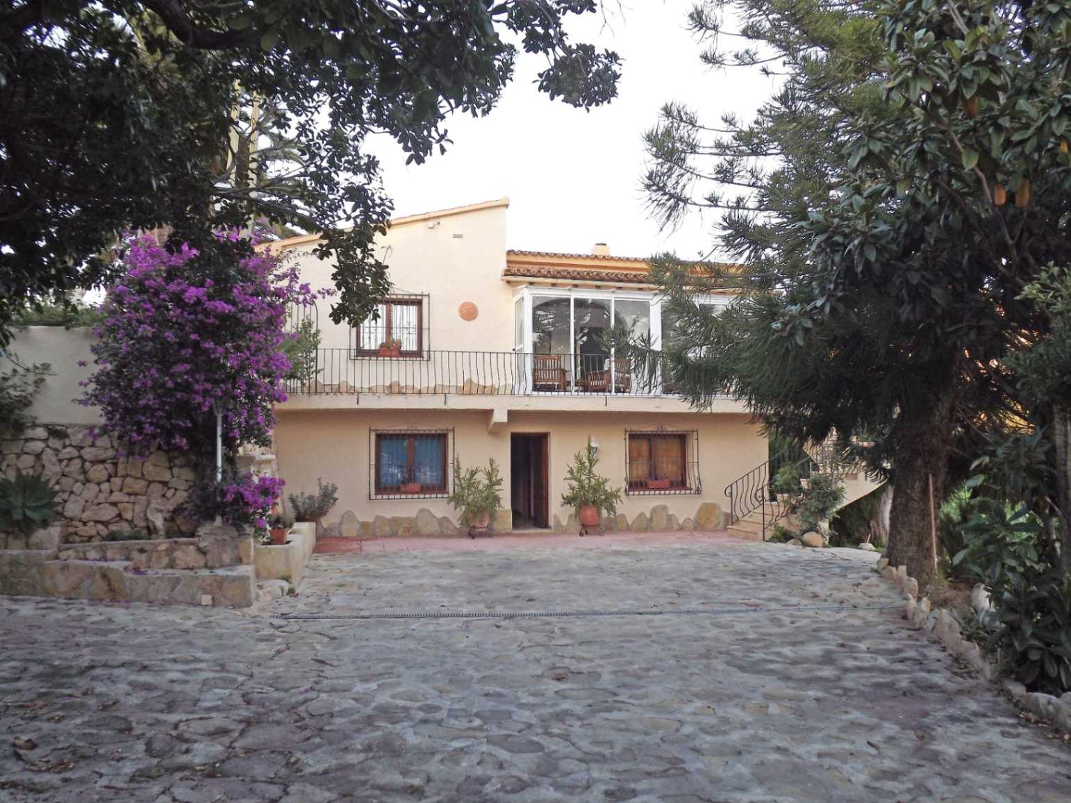 Fachada principal de la villa con entrada empedrada, terraza acristalada en la planta superior y detalles de piedra natural rodeados de vegetación mediterránea.