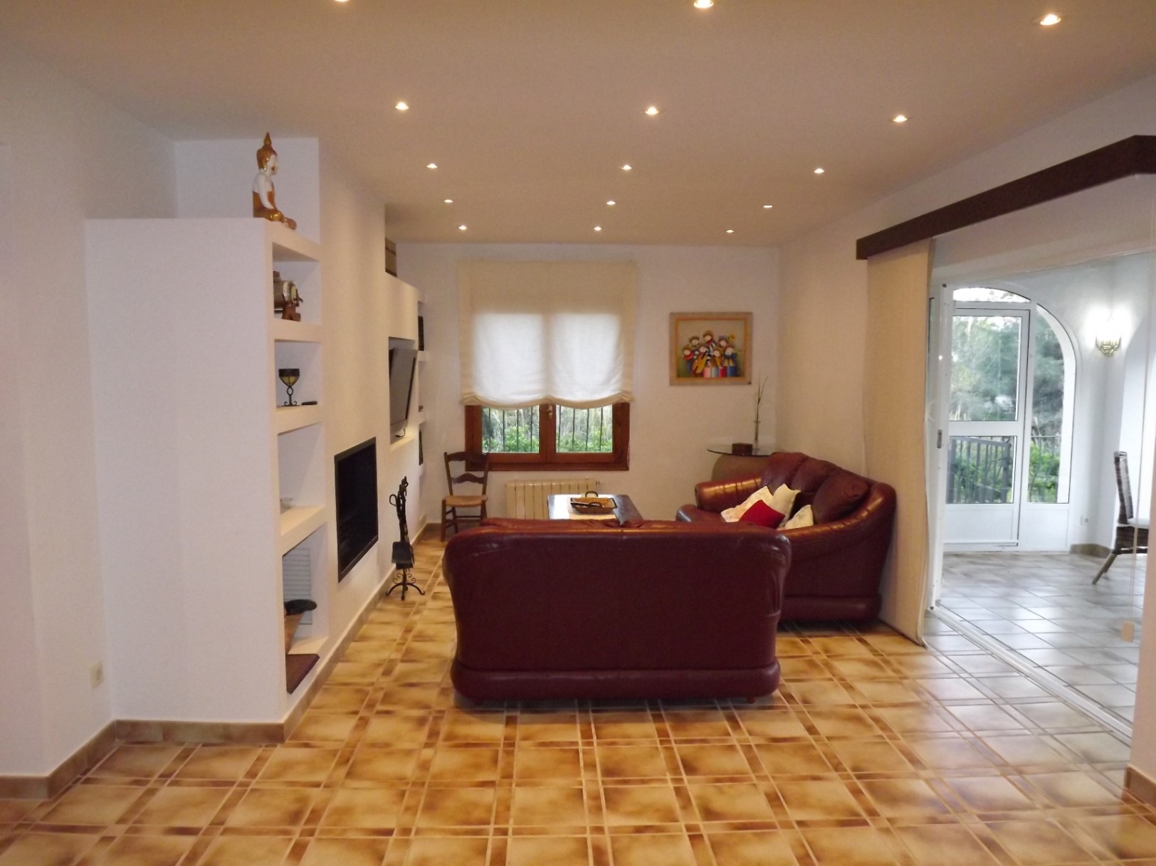 Salón con sofás de cuero granate, chimenea integrada y estantería de obra. El suelo de gres rústico y la iluminación empotrada conectan con la terraza acristalada.