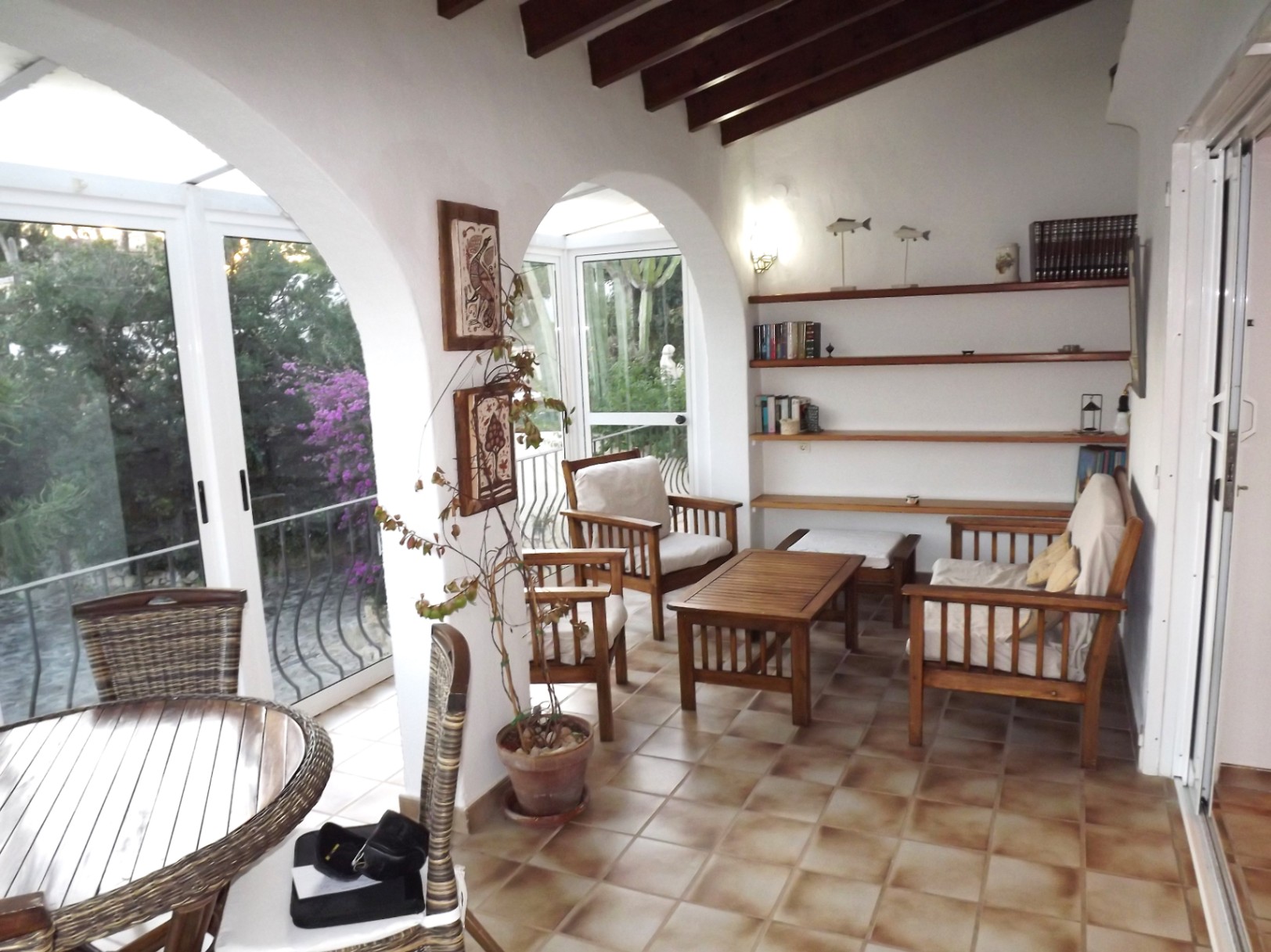 Terraza acristalada con arcos tradicionales, techos de vigas de madera y suelos de terracota. Incluye mobiliario de madera, estanterías integradas y vistas al jardín.