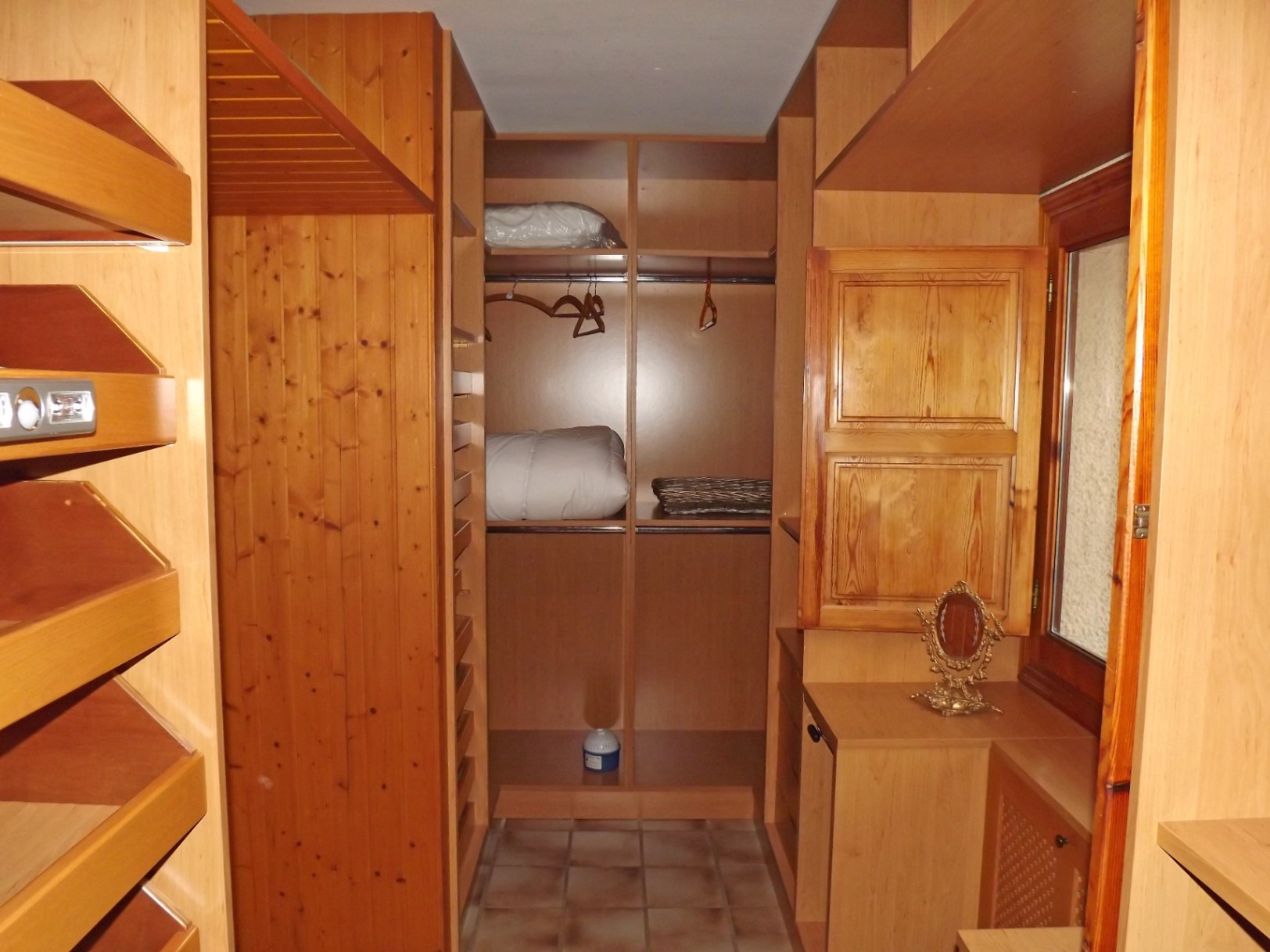 Vestidor revestido íntegramente en madera con estanterías a medida, barras para colgar y suelos de baldosa. Incluye una ventana con contraventana de madera y zona de tocador.