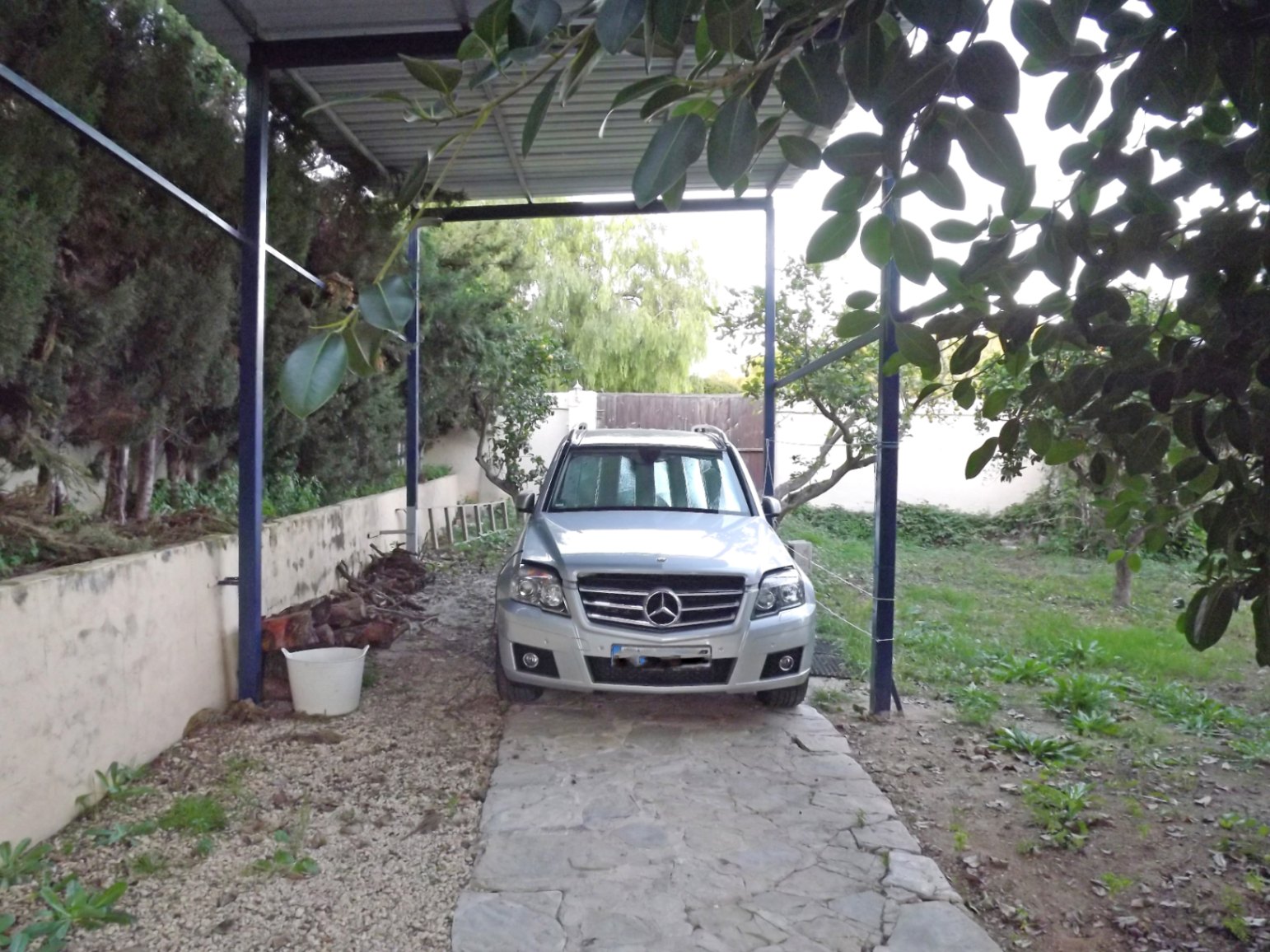 Zona de aparcamiento cubierta con estructura metálica y suelo de piedra natural, rodeada de vegetación y árboles frutales en la amplia parcela.