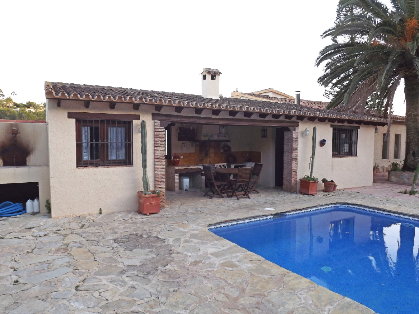 Zona exterior con piscina de azulejos azules, amplia terraza de piedra natural y porche cubierto con vigas de madera y barbacoa integrada.