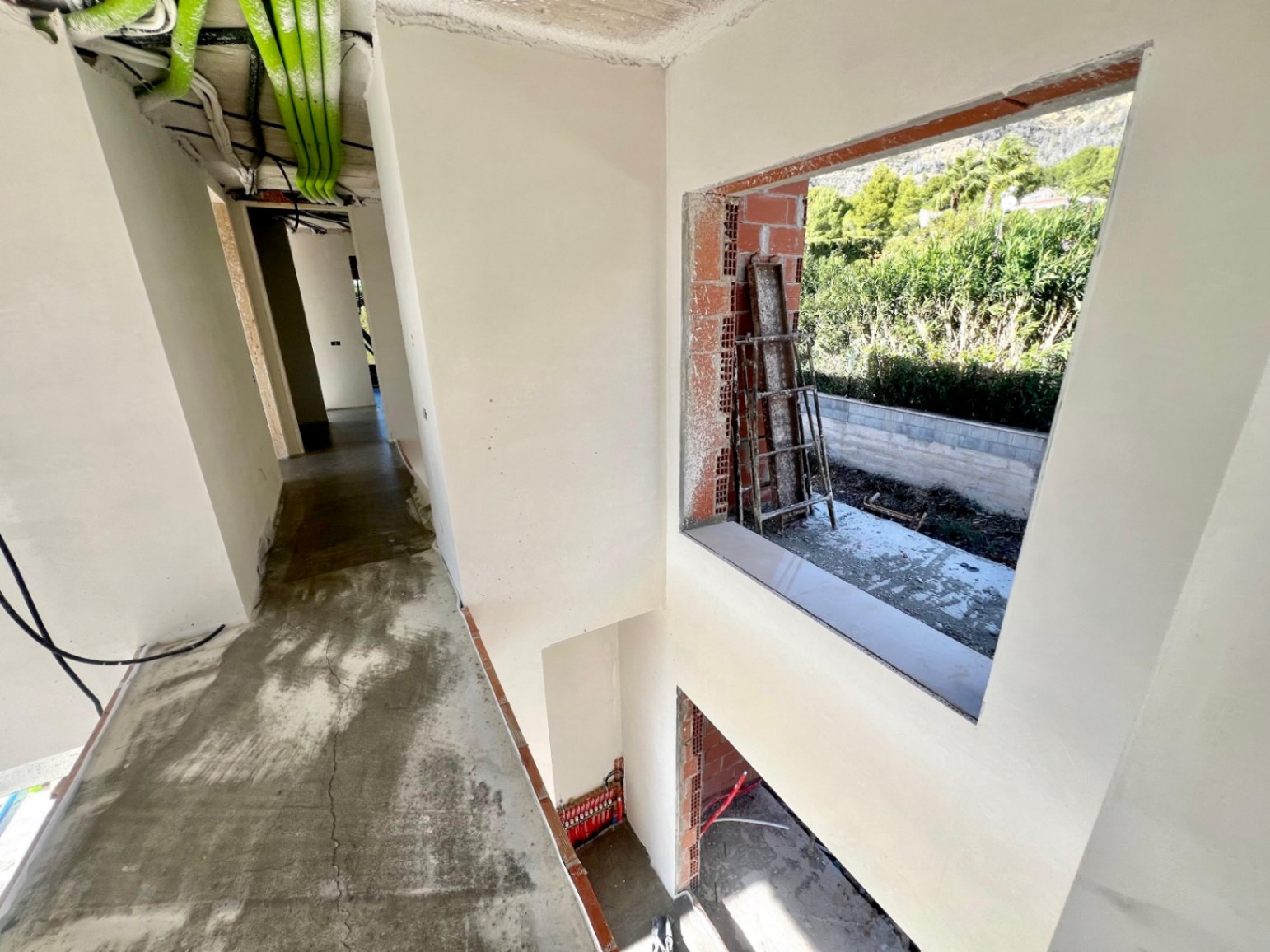 Pasillo interior en fase de construcción con techos de doble altura, instalaciones técnicas visibles y un amplio ventanal hacia el entorno natural.