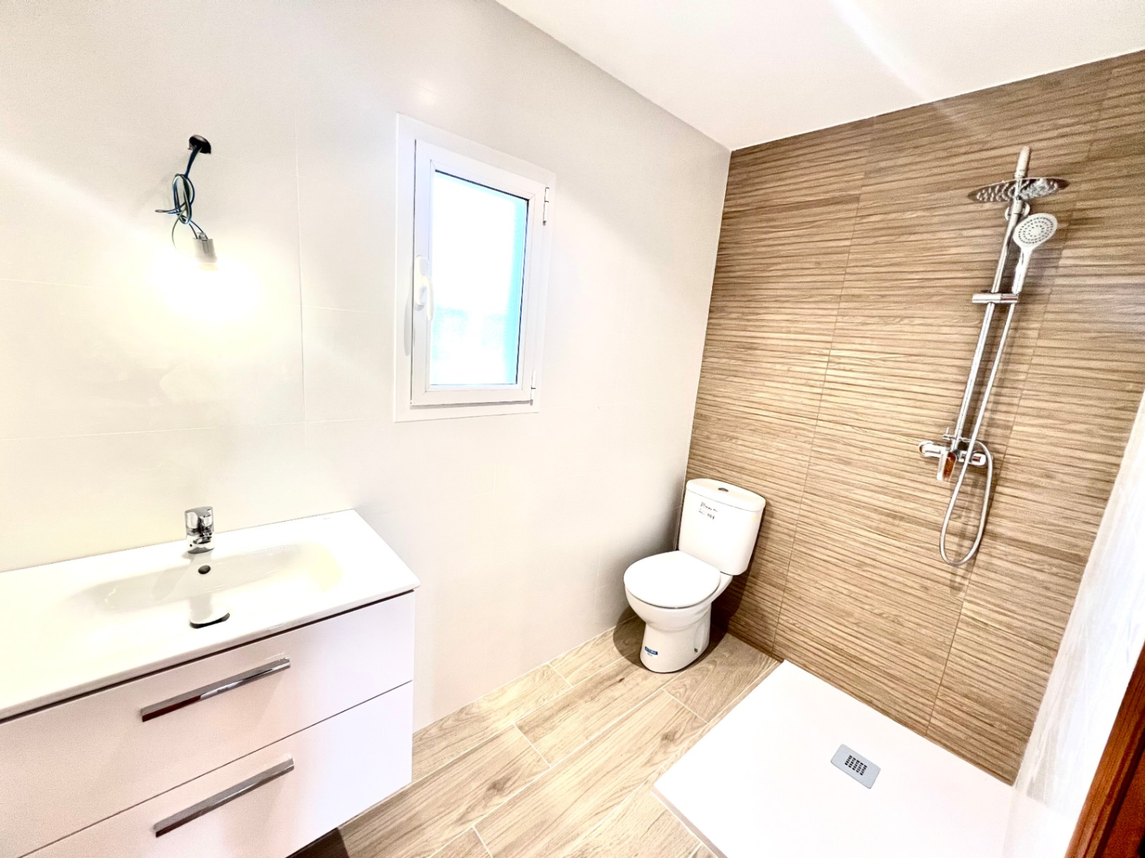 Baño moderno con plato de ducha, grifería de efecto lluvia y paredes revestidas en tonos madera y blanco. Incluye mueble de lavabo suspendido y ventana de PVC.