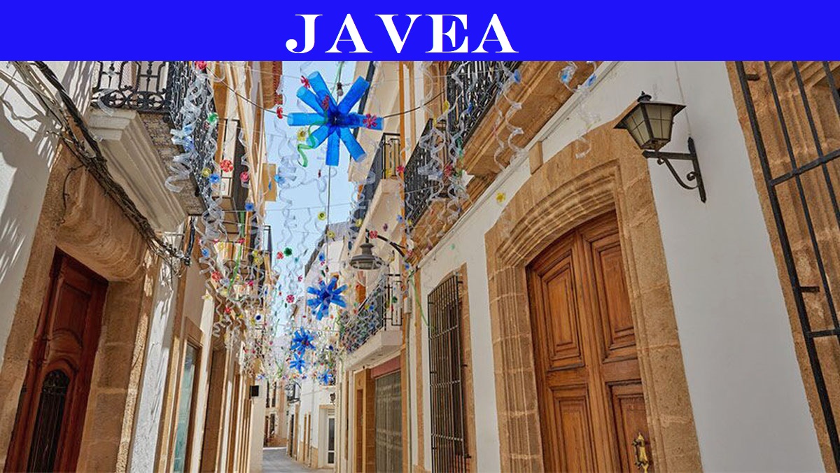 Calle tradicional en el casco antiguo de Jávea con fachadas blancas, marcos de piedra tosca y coloridas decoraciones festivas suspendidas.