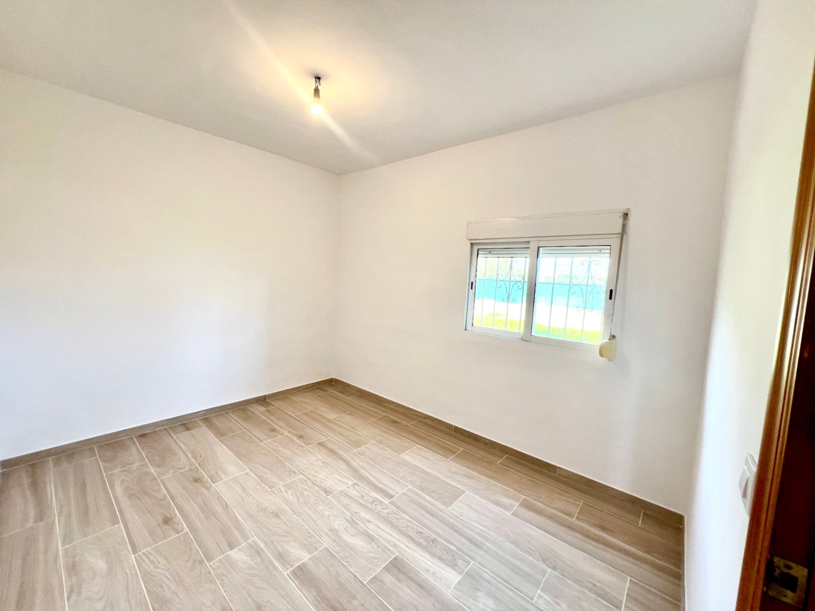 Dormitorio luminoso con suelos de gres efecto madera, paredes blancas lisas y ventana con reja de seguridad y persiana integrada.