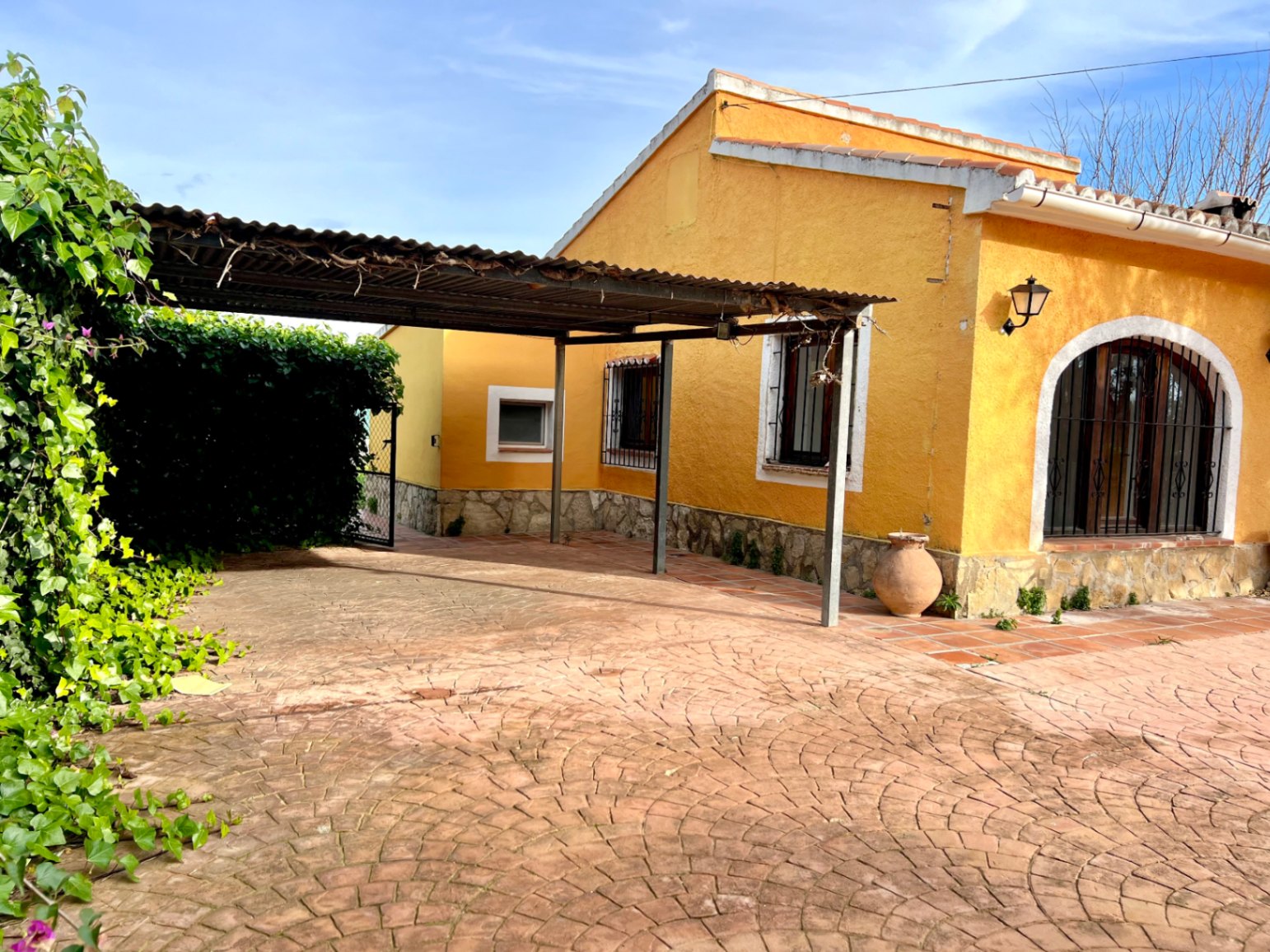 Fachada amarilla con zócalo de piedra, zona de aparcamiento cubierto por pérgola metálica y suelo de hormigón impreso con diseño de abanico.