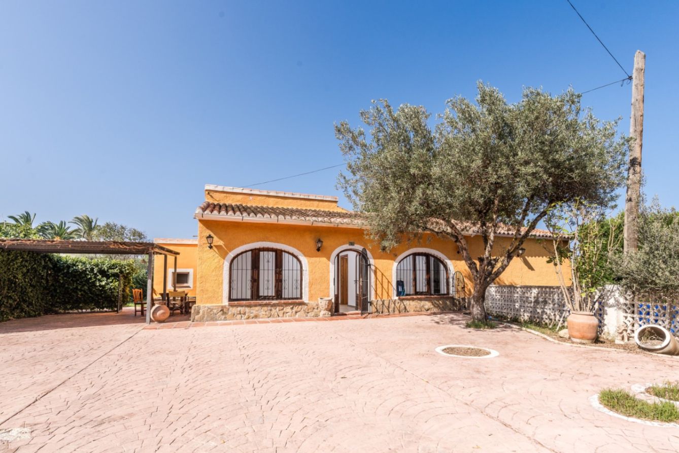 Fachada de color ocre con ventanas de arco y zócalo de piedra. El exterior cuenta con un amplio patio pavimentado, un olivo maduro y una zona de pérgola lateral.