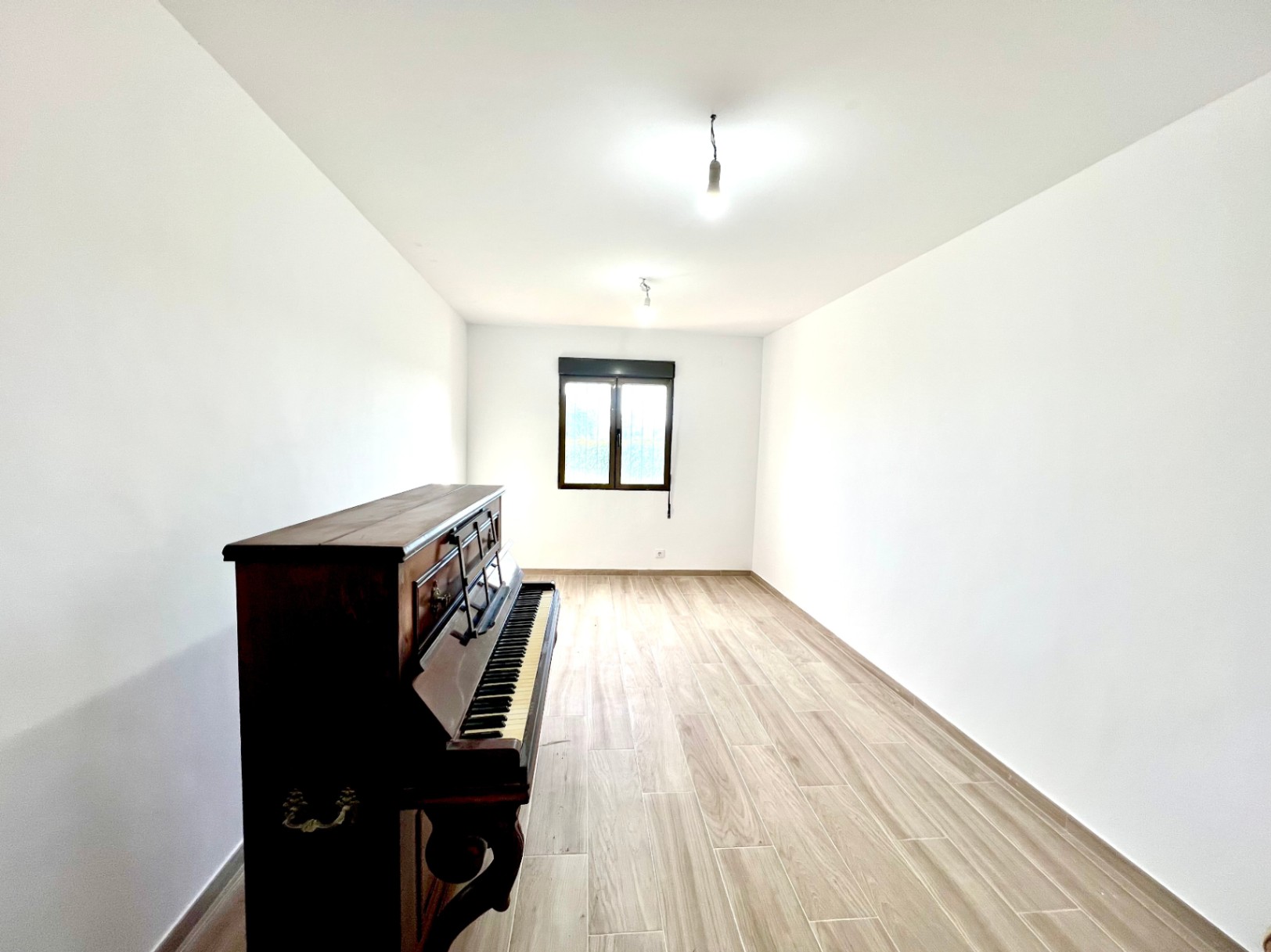 Habitación alargada con suelos de gres efecto madera, paredes blancas y un piano vertical clásico. Incluye una ventana de marco oscuro que aporta luz natural.