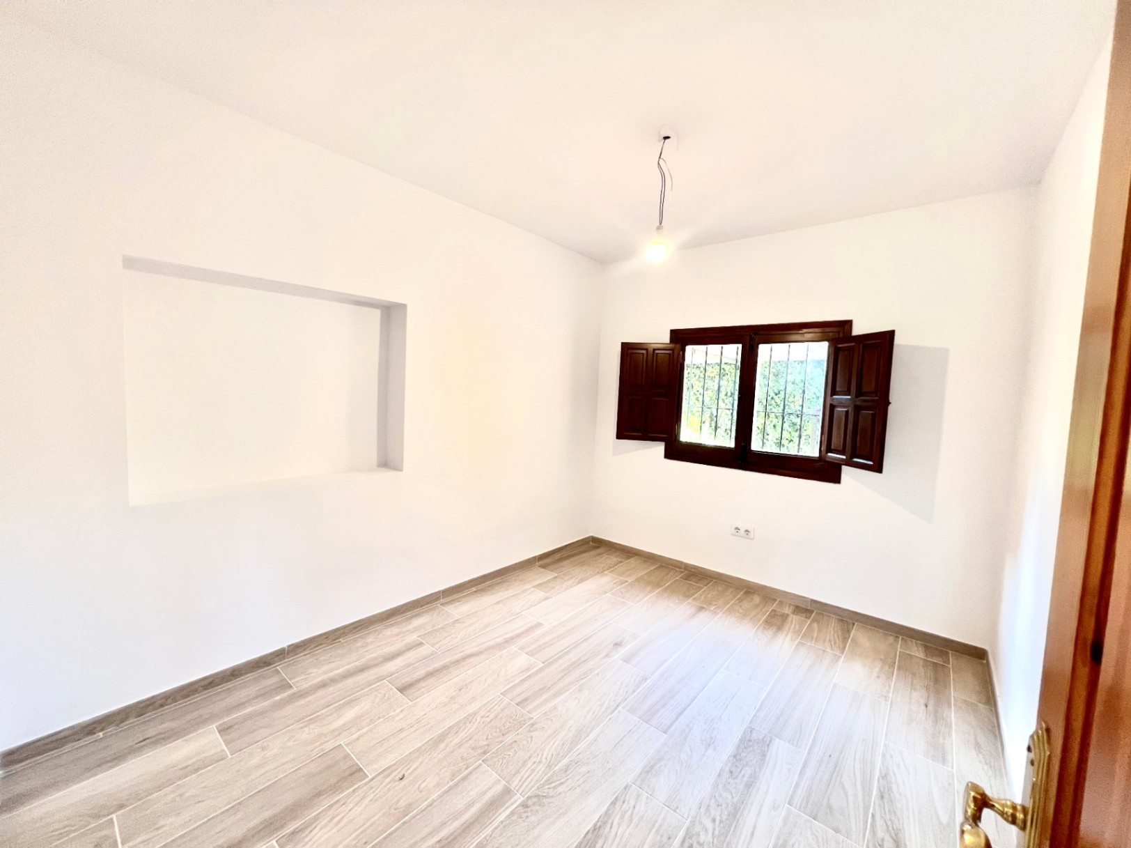 Habitación luminosa con suelos de gres efecto madera, paredes blancas, un nicho decorativo integrado y ventana con contraventanas de madera oscura.