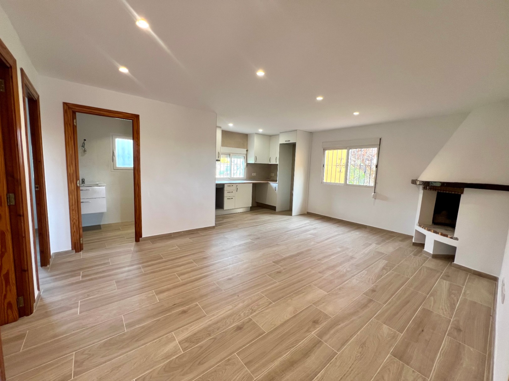 Salón comedor diáfano con chimenea de rincón, suelos de gres efecto madera y cocina abierta. Incluye iluminación por focos empotrados y marcos de puertas en madera natural.