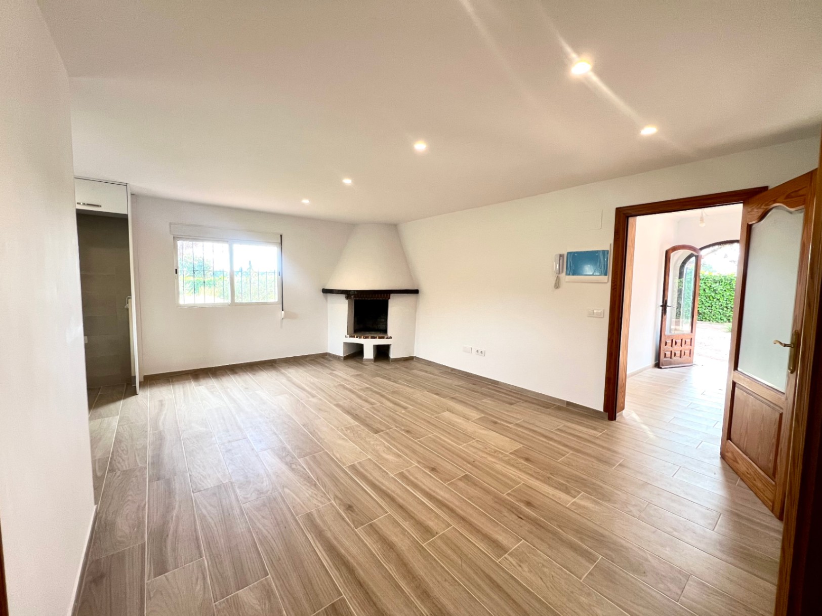 Salón diáfano con chimenea de rincón, suelos de gres efecto madera y focos empotrados. La estancia conecta con el recibidor y ofrece vistas al jardín exterior.