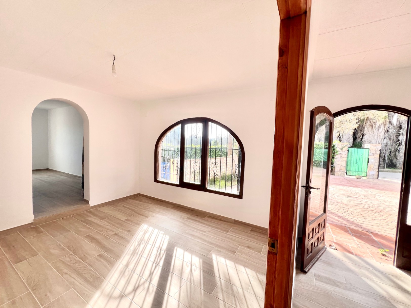 Salón luminoso con suelos cerámicos imitación madera, ventanas de arco en madera oscura y acceso directo al patio exterior pavimentado.