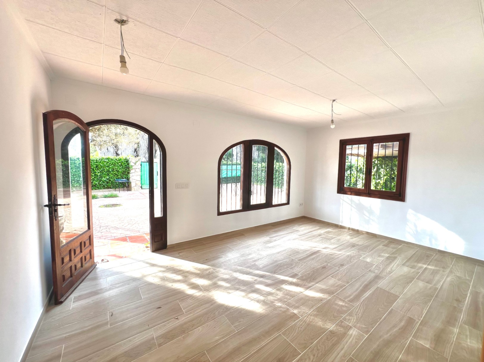 Salón luminoso con suelos de gres efecto madera, carpintería de madera oscura en ventanas de arco y acceso directo al patio exterior.