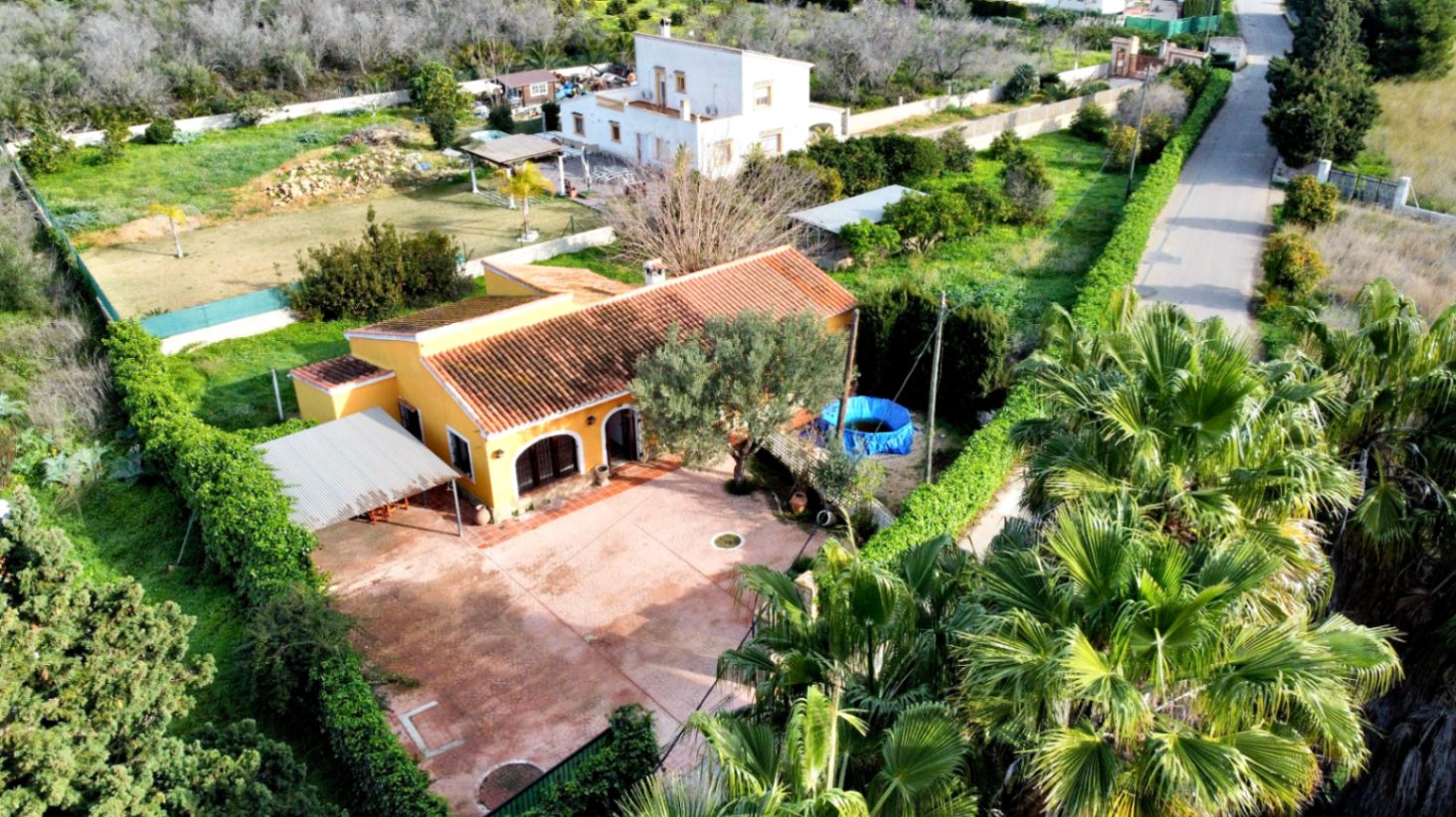 Vista aérea de la propiedad que muestra la fachada amarilla, el amplio patio pavimentado, el porche arqueado y la zona de aparcamiento cubierto rodeada de vegetación.
